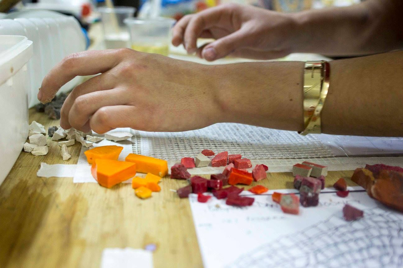Hands sorting cut tile pieces by colour on a studio workbench